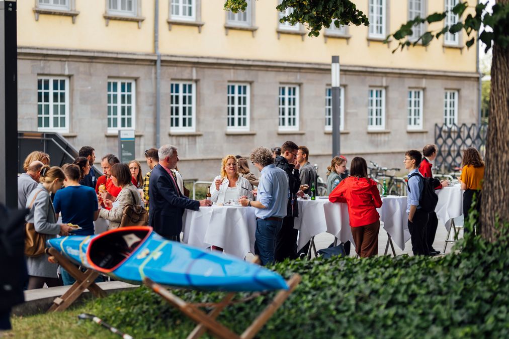zur Vergrößerungsansicht des Bildes: im Vordergrund steht das hellblaue Kajak einer Stipendiatin, im Hintergrund stehen die Gäste an Stehtischen und essen, trinken und unterhalten sich