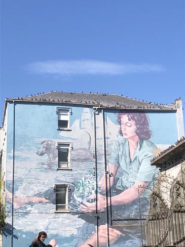 Eine Hauswand komplett mit einem Gemälde bedeckt. Zu sehen sind eine Frau, die eine Zimmerpflanze in den Händen hält und im Wasser sitzt. Im Hintergrund steht ein Dackel. Dahinter sieht man ein Segelboot. Unten links im Bildrand sitzt eine weitere Person.