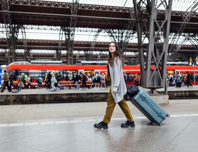 Colour photo: A person walks with a suitcase along the station track. There is a train and many travellers in the background