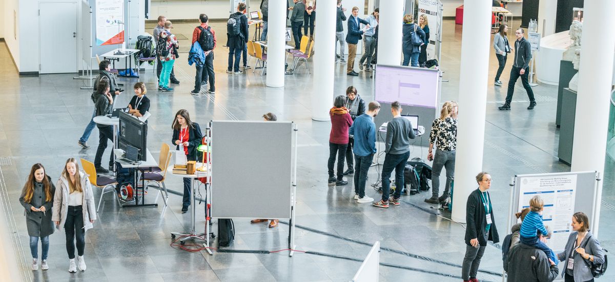 Vogelperspektive in das Foyer des Neuen Augusteum. Mehrere Menschengruppen stehen an Informationstafeln und lesen oder unterhalten sich.