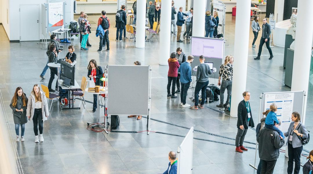 Vogelperspektive in das Foyer des Neuen Augusteum. Mehrere Menschengruppen stehen an Informationstafeln und lesen oder unterhalten sich.