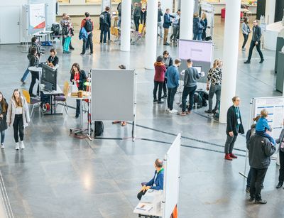 Vogelperspektive in das Foyer des Neuen Augusteum. Mehrere Menschengruppen stehen an Informationstafeln und lesen oder unterhalten sich.