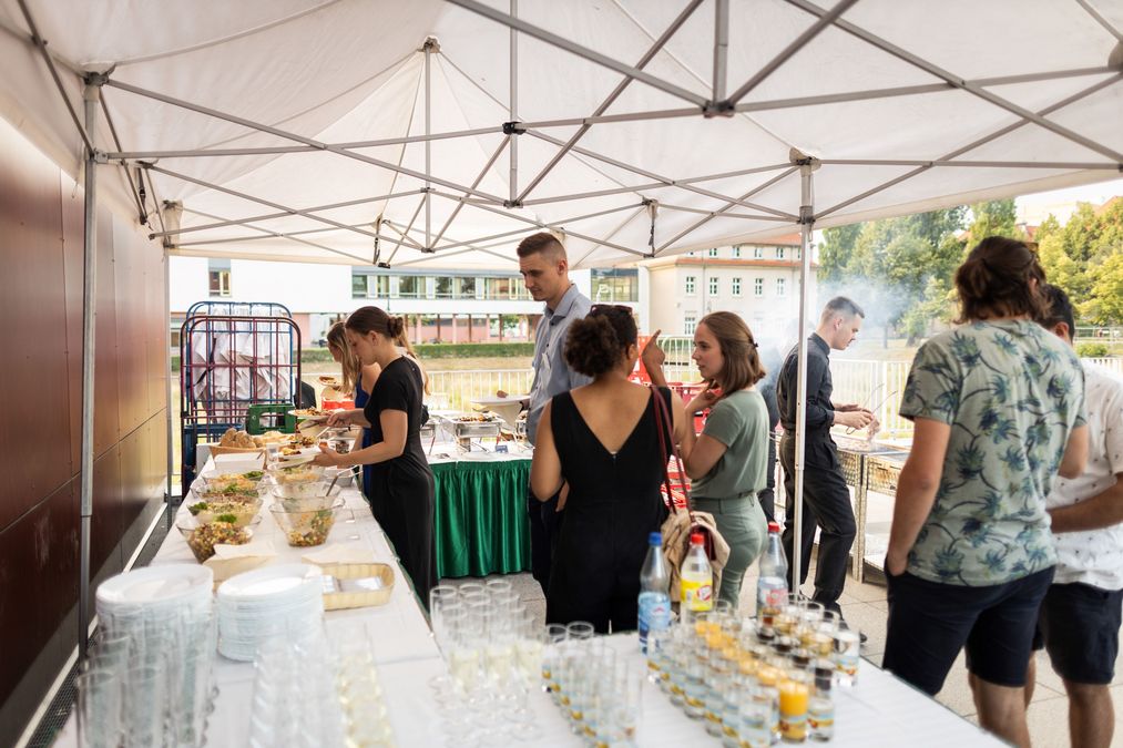 zur Vergrößerungsansicht des Bildes: Farbfoto: Buffet im Vordergrund stehen Getränke, an der Seite sind verschiedene Salate, im Hintergrund wird gegrillt