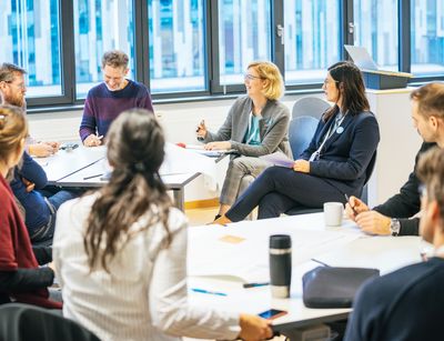 Farbfoto: Acht Personen unterschiedlichen Geschlechts sitzen in einer Runde am Tisch und diskutieren miteinander.