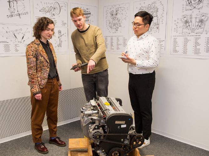 Drei Personen stehen um einen Motor herum, im Hintergrund hängen technische Zeichnungen an den Wänden. Einer der zwei Männer zeigt auf den Motor, die Frau hört ihm zu. Der zweite Mann macht sich handschriftlich Notizen. Foto: Bodo Tiedemann