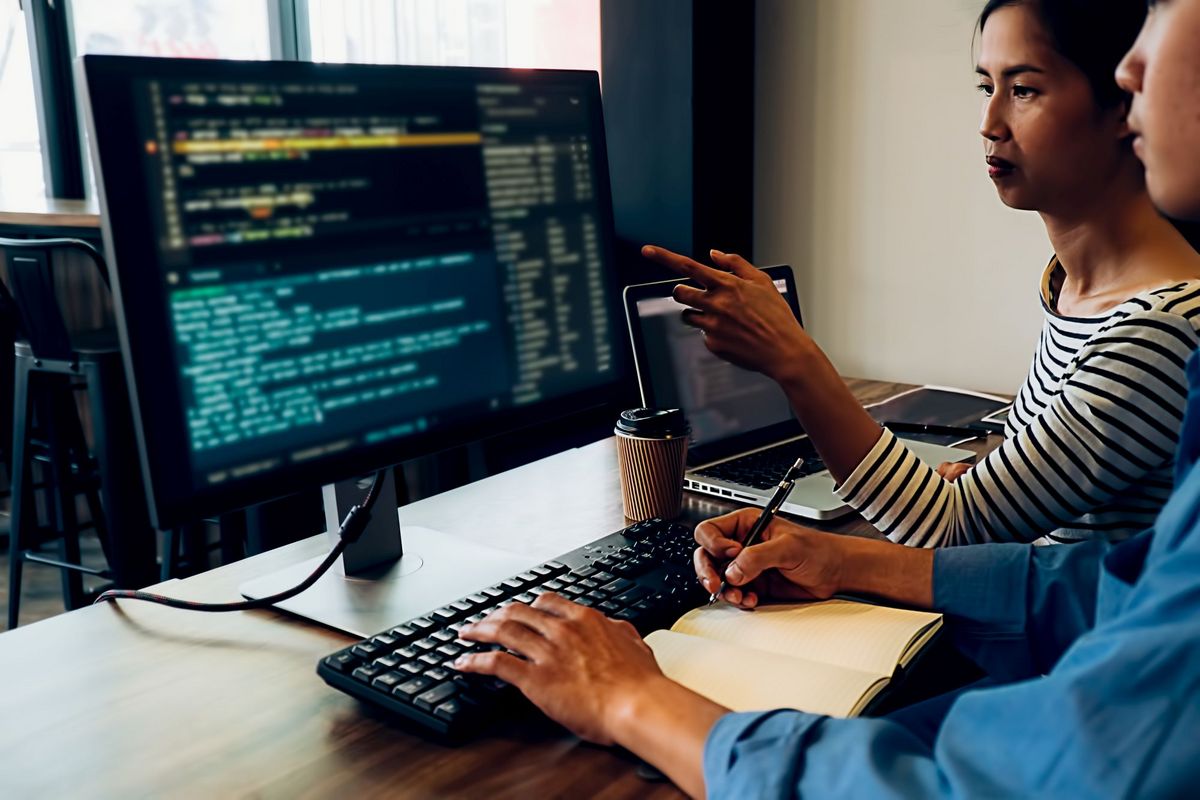 zur Vergrößerungsansicht des Bildes: Farbfoto: Ein Mann und eine Frau sitzen an einem Computerarbeitsplatz und blicken auf einen Monitor, der Programmiercode anzeigt. Die Frau zeigt auf den Monitot. Der Mann tippt mit der linken Hand und macht sich mit der rechten Hand Notizen in einem Buch.