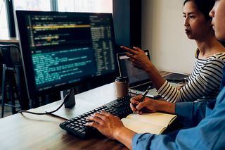 Farbfoto: Ein Mann und eine Frau sitzen an einem Computerarbeitsplatz und blicken auf einen Monitor, der Programmiercode anzeigt. Die Frau zeigt auf den Monitot. Der Mann tippt mit der linken Hand und macht sich mit der rechten Hand Notizen in einem Buch.