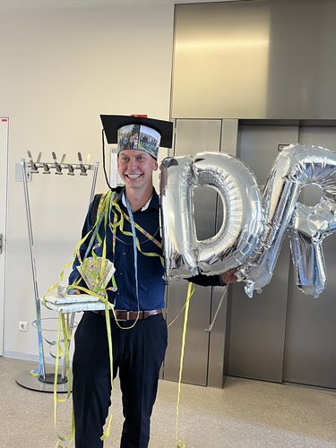 A smiling man standing in front of the elevator with a doctoral hat on his head and baloons and presents in his hands
