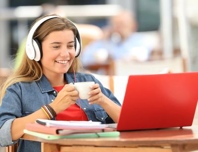 Studentin sitzt vor ihrem roten Laptop mit einem Kaffee und hat weiße Kopfhörer auf, Foto: Colourbox