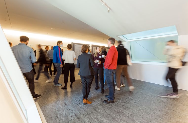 People stand in groups of two and three and talk, while other individually explore posters displaying scientific data