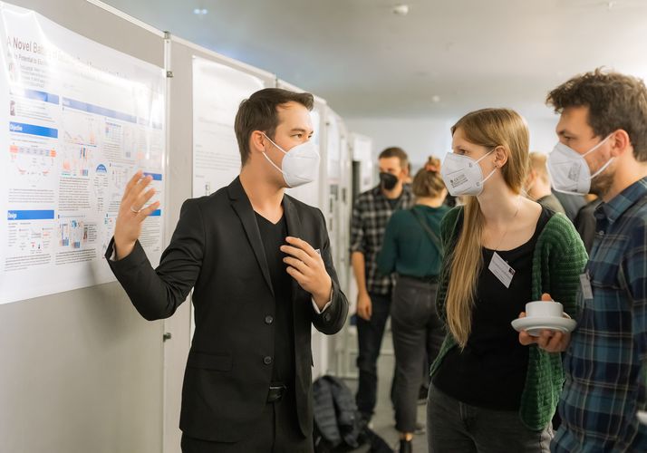 A postdoc stands in front of the poster wall explaining his data to two doctoral candidadtes