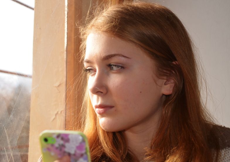 Junge Frau blickt mit Handy in der Hand durchs Fenster