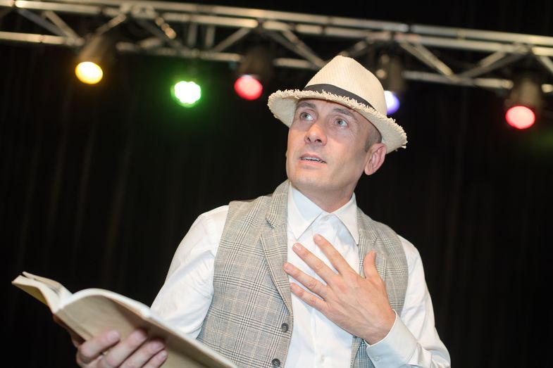Ein älterer männlicher Schauspieler mit Hut steht auf einer Bühne und trägt etwas vor. In seiner Hand hält er ein aufgeschlagenes Buch.