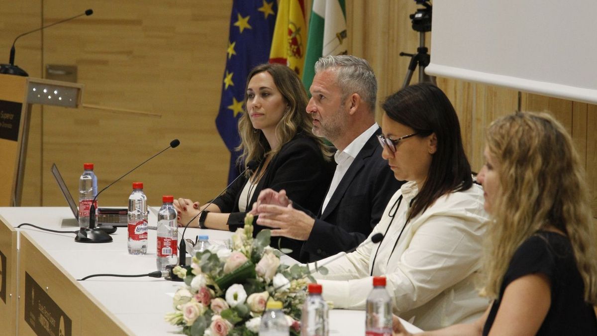 enlarge the image: 4 people sitting on a stage at a table facing the audience, photographed from the side