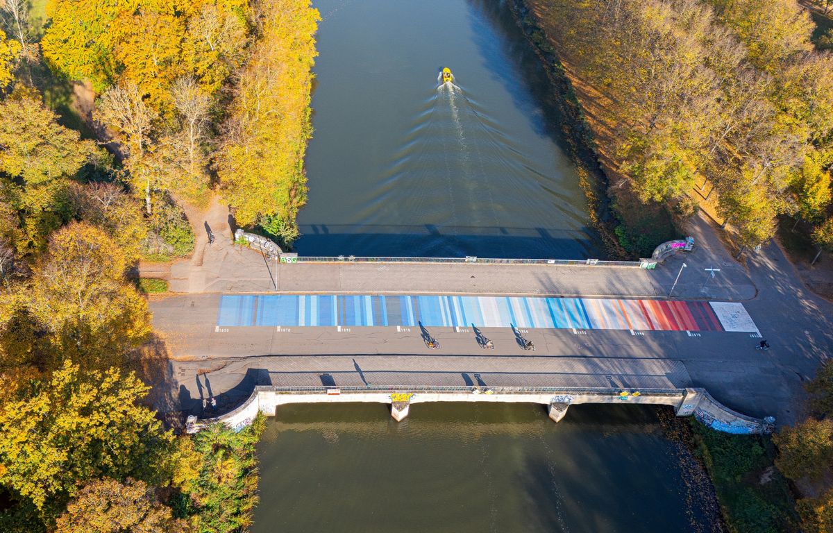 enlarge the image: Luftaufnahme: Vogelperspektive auf die Sachsenbrücke, auf der Farbstreifen von Blau bis Rot den zeitlichen Verlauf der Erderwärmung abbilden.