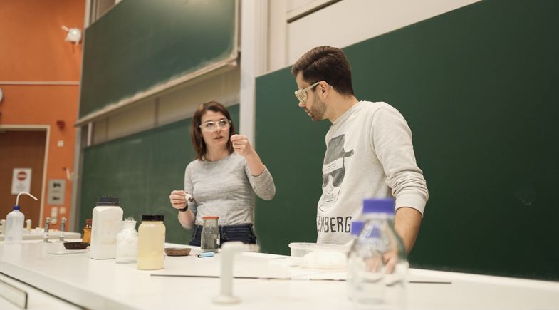 Marika Kandler und Jack Pop stehen am Experimentiertisch in einem Hörsaal.