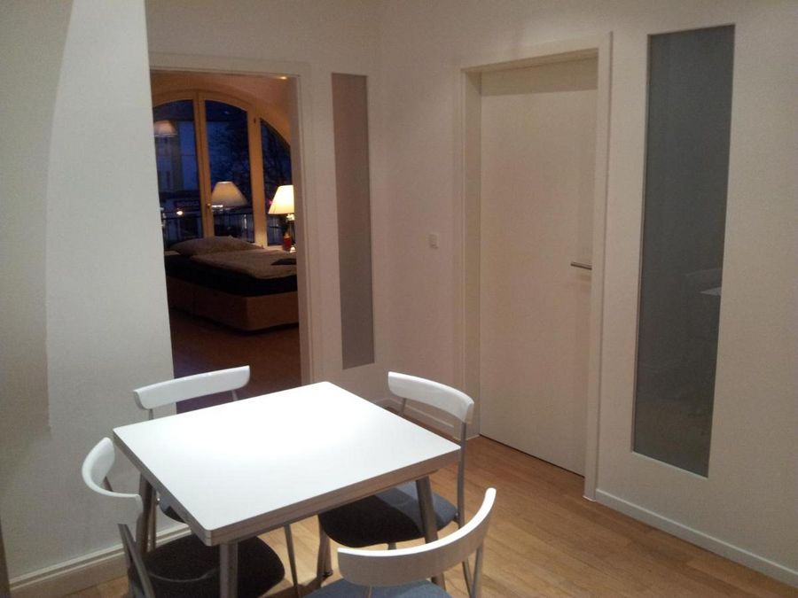 enlarge the image: Farbfoto: Innenaufnahme eines Wohnraums mit Essecke. Ein quadratischer Tisch mit vier weißen Stühlen steht im Mittelpunkt. Im Hintergrund kann man ins Schlafzimmer blicken