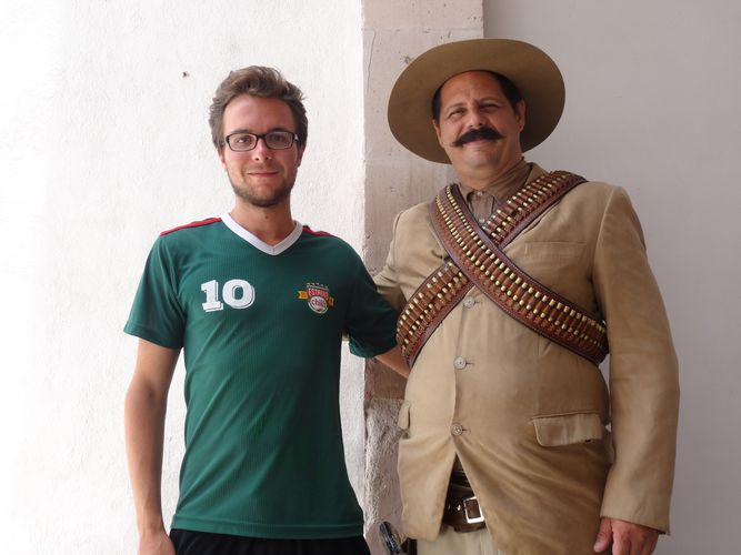 Zwei Männer stehen vor einer weißen Wand. Der Jüngere trägt ein grünes Fußballtrikot, während der Ältere ein Kostüm trägt. Er ist als Mexikaner verkleidet. Über seinem braunen Oberteil prangert ein Patronengurt.