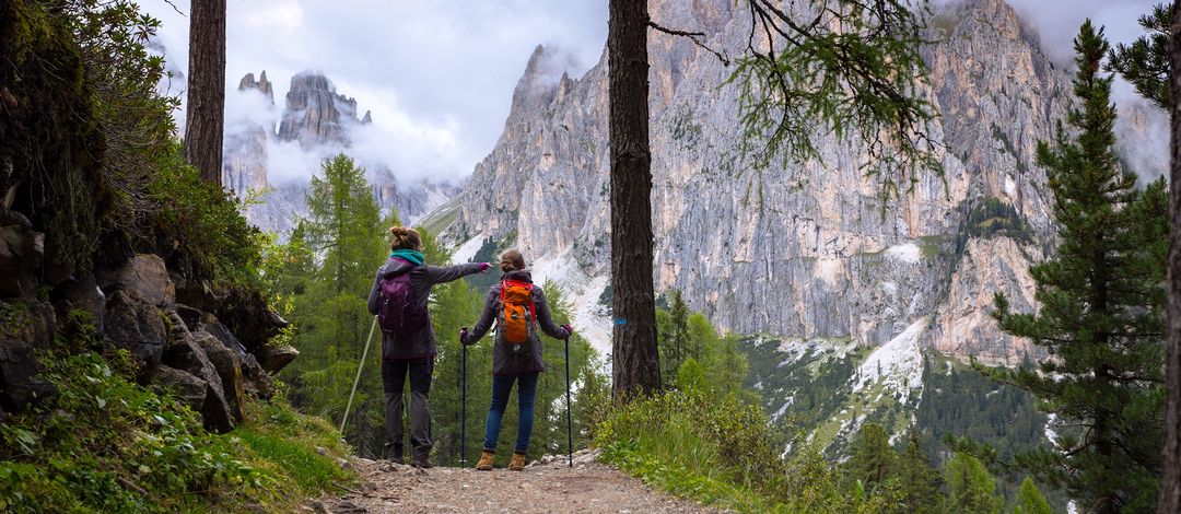 Zwei junge Frauen in Wanderkleidung befinden sich in einer Berglandschaft. Ihr Blick ist in Richtung des Berggipfels gerichtet. Eine der Frau deutet mit ihrem Finger in Richtung Gipfel.