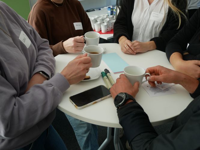 Das Bild zeigt fünf Menschen, die an einem runden Stehtisch bei Kaffee zusammenkommen und sich austauschen. Ihre Gesichter sind auf dem Bild nicht zu erkennen.