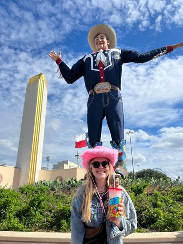 Person steht mit pinken Hut und Sonnenbrille vor einer Cowboy-Statue bei Tag