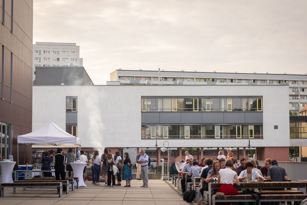 zur Vergrößerungsansicht des Bildes: Farbfoto: Förder:innen und Stipendiat:innen stehen und sitzen auf einer Terrasse, im Hintergrund wird gegrillt