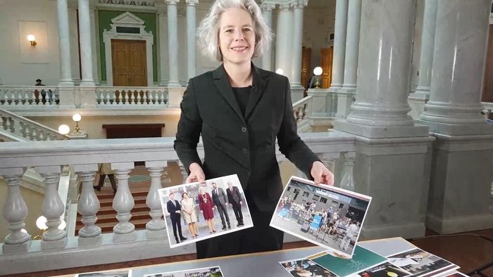 Die Rektorin steht an einem Tisch auf dem viele Bilder von Ereignissen aus dem ersten Amtsjahr ausgelegt sind. Sie schaut in die Kamera und hält zwei dieser Bilder in der Hand. Im Hintergrund erkennt man die Räumlichkeiten der Universitätsbibliothek.
