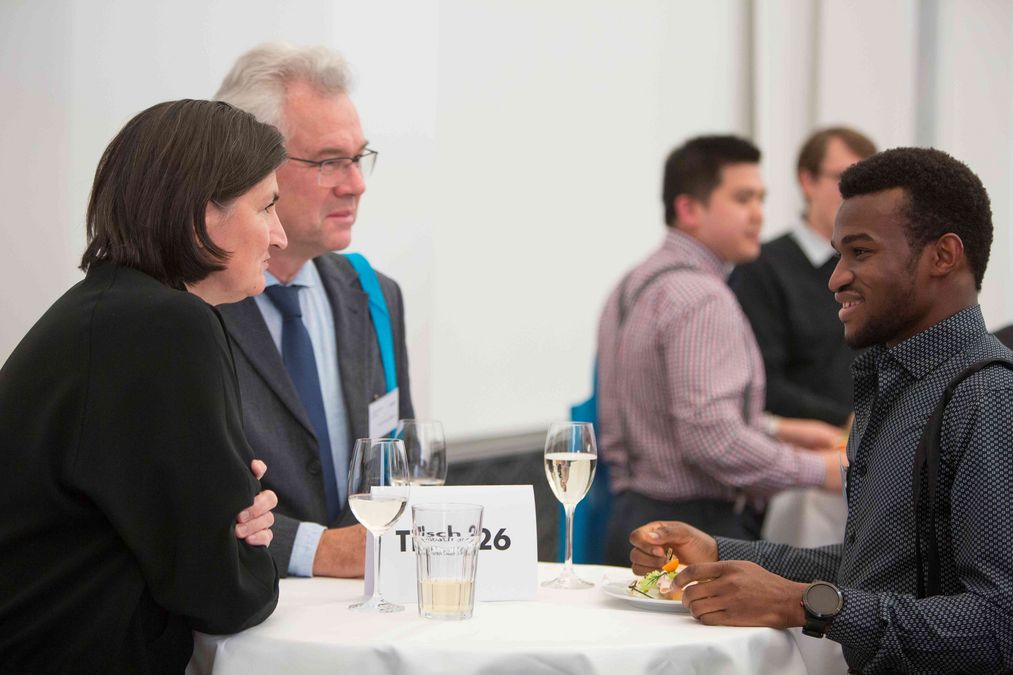 zur Vergrößerungsansicht des Bildes: Professor Ingo Dähnert und seine Frau stehen mit ihrem Stipendiaten an einem Stehtisch und unterhalten sich, auf dem Tisch stehen Gläser