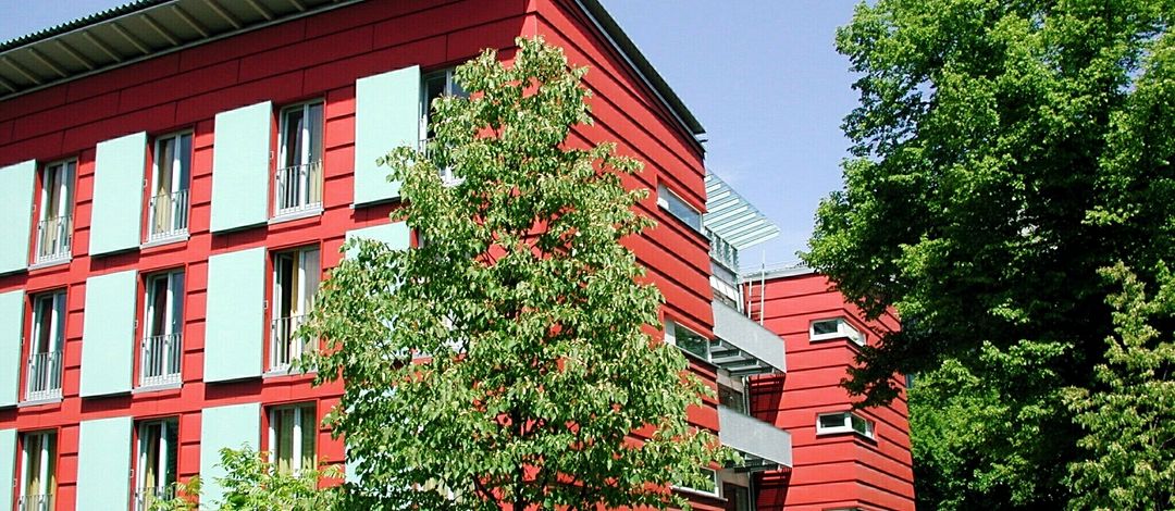 Rote Fassade des Werner-Heisenberg-Hauses der Universität. Die Bäume vor dem Gebäude lassen erahnen, dass es in einer besonders grünen Umgebung steht.