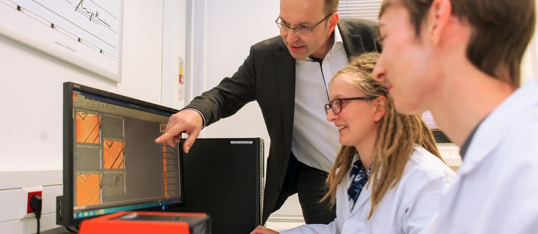 Foto: zwei Studierende im Arztkittel sitzen vor einem PC während ein Professor ihnen etwas erklärt