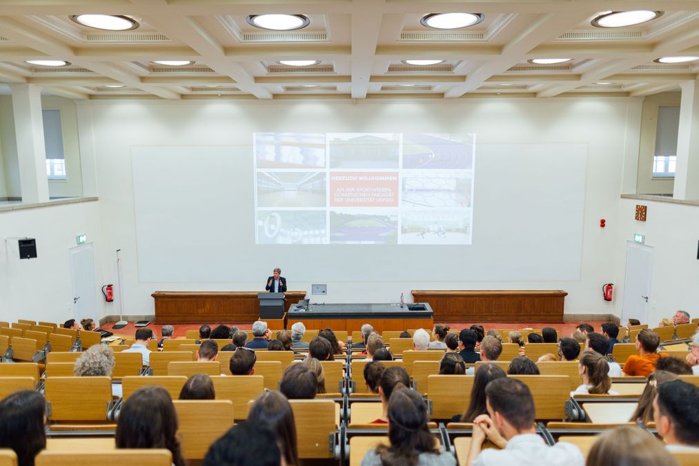 zur Vergrößerungsansicht des Bildes: Fördernde und Studierende sitzen in Hörsaal, im Hintergrund steht Professor Hovemann und begrüßt die Gäste an der Sportwissenschaftlichen Fakultät
