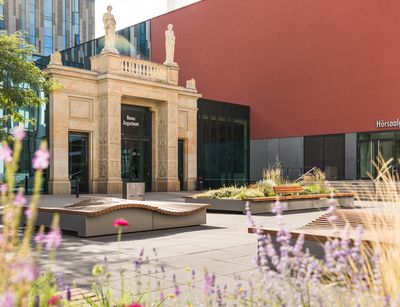 Architekturfotografie: historischer Torbogen aus braunem Stein, das Bild ist im Sommer aufgenommen. Es sind keine Menschen auf dem Campus zu sehen.