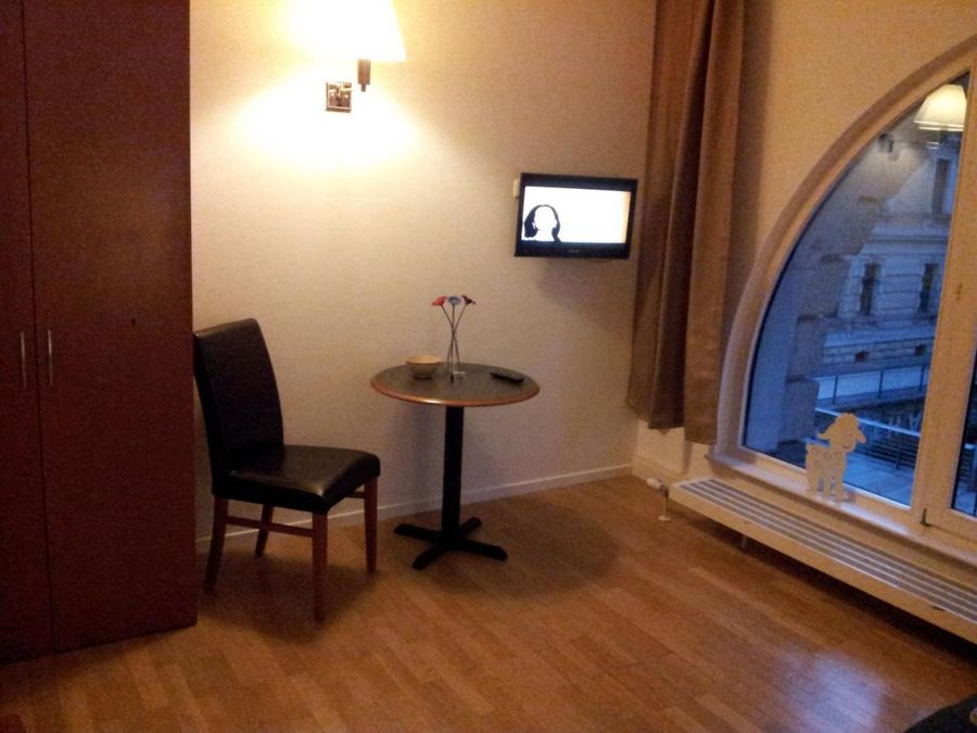 enlarge the image: Farbfoto: Innenansicht eines Wohnraums mit einem Stuhl, einem kleinen runden Tisch, einem kleinen Fernseher an der Wand und einem bodentiefen Fenster