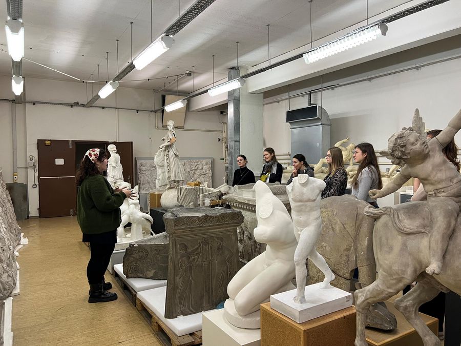 zur Vergrößerungsansicht des Bildes: im Vordergrund sind verschiedene Statuen und historische Gipsabgüsse griechischer und römischer Skulpturen, links steht eine Studierende, die die Gäste, im rechten Hintergrund stehend, durch die Sammlung führt