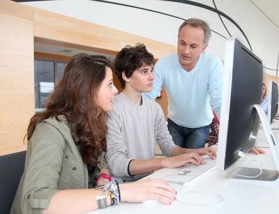 Ein junges Mädchen und ein junger Mann sitzen gemeinsam an einem Schreibtisch an einem Computer. Rechts neben Ihnen steht ein älterer Mann und beugt sich zu ihnen und erklärt. Das Mädchen schaut zum Lehrer, der Junge blickt in den Monitor.