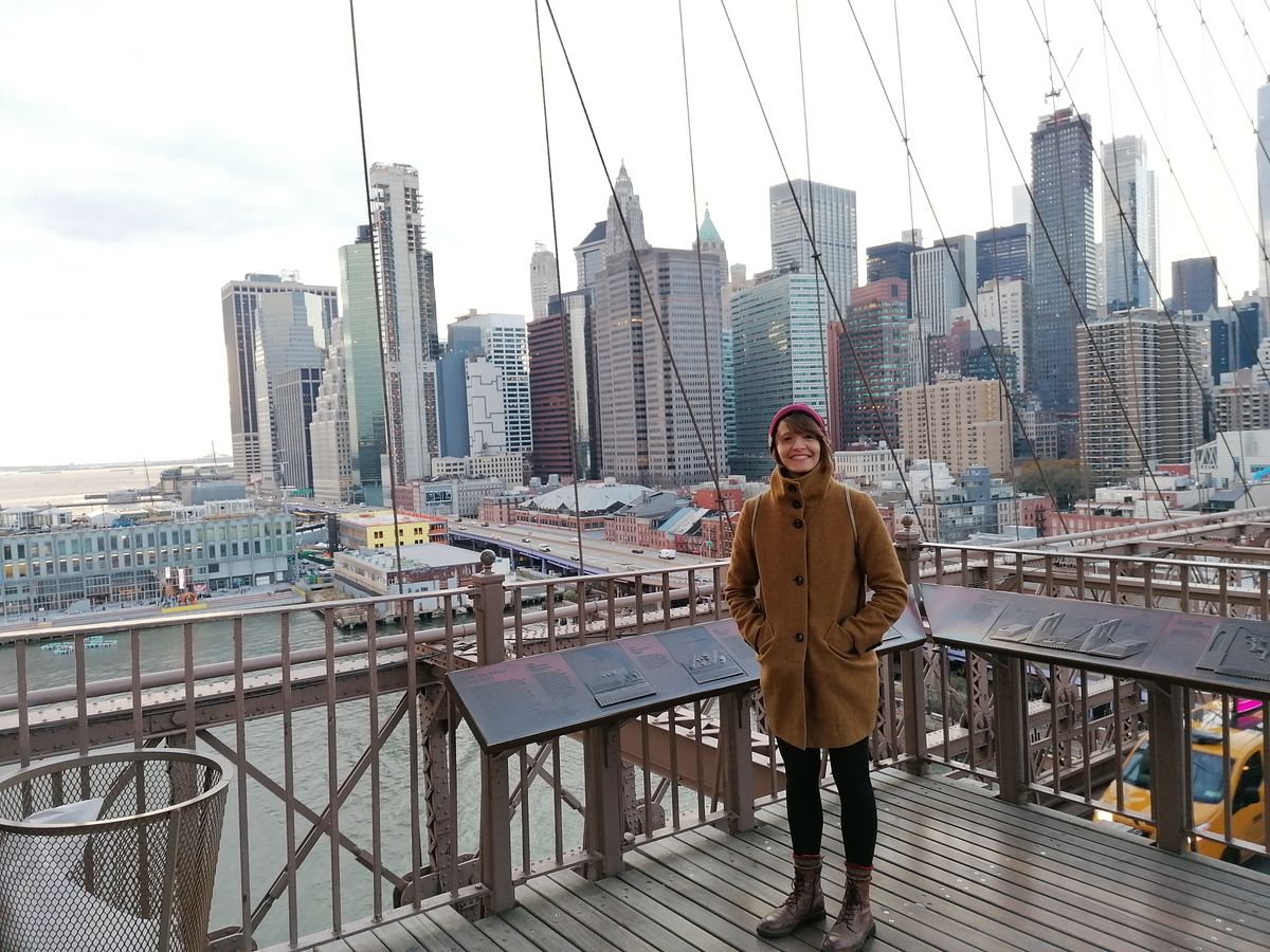 enlarge the image: A woman standing on Brooklyn Brigde with the skyline in the background.