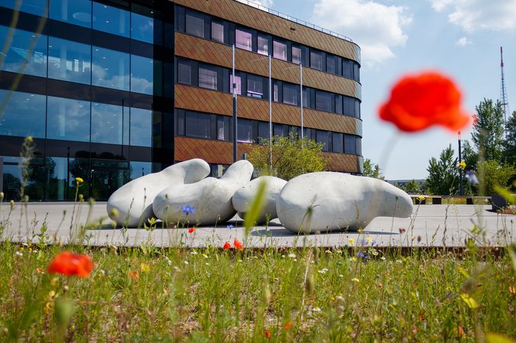 Farbfoto: Im Hintergrund steht ein mehrstöckiger, moderner Bürokomplex, davor ist eine Kunstinstallation in Form von großen "Betoklecksen", im Vordergrund ist eine Wiese mit roten Mohnblumen zu sehenn