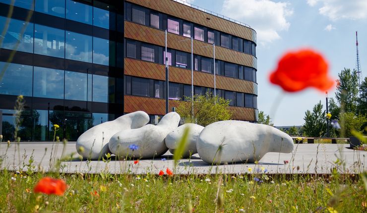 Farbfoto: Im Hintergrund steht ein mehrstöckiger, moderner Bürokomplex, davor ist eine Kunstinstallation in Form von großen "Betoklecksen", im Vordergrund ist eine Wiese mit roten Mohnblumen zu sehenn
