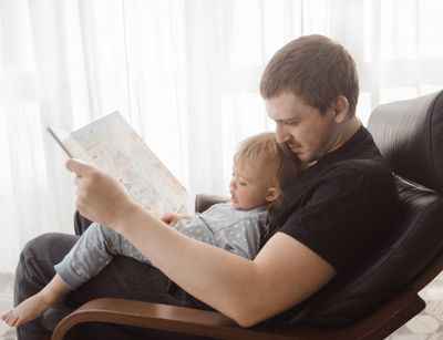 Zu sehen ist ein junger Mann mit einem Kleinkind auf dem Schoß. Sie schauen gemeinsam ein Buch an.