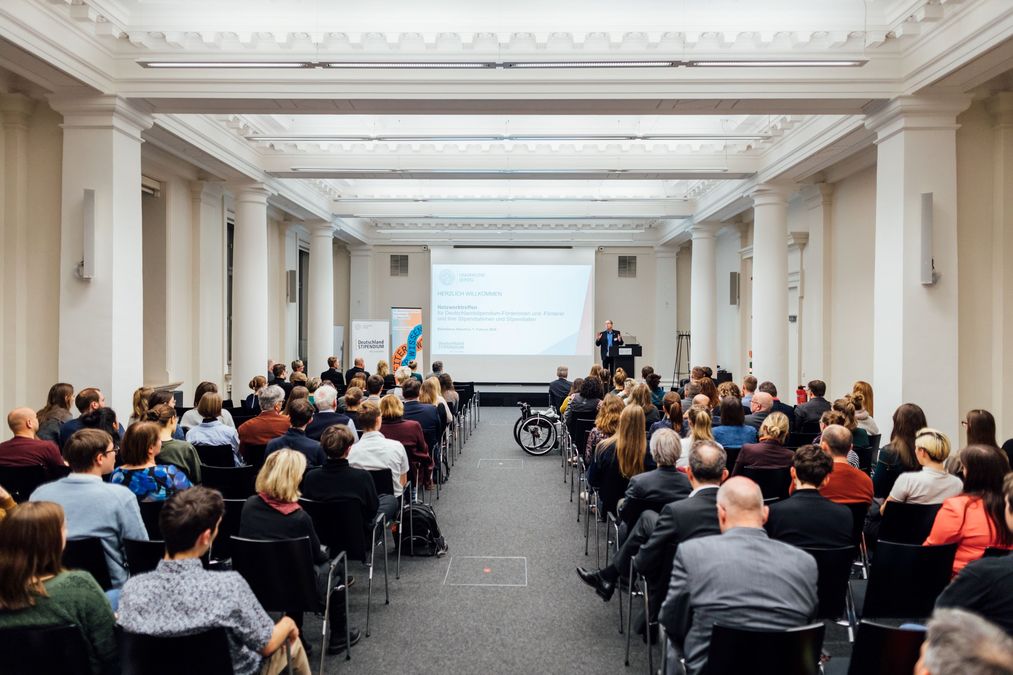 zur Vergrößerungsansicht des Bildes: Prorektor Professor Gläser steht auf einer Bühne vor den Gästen, die in Reihen im Vortragsraum der Albertina sitzen und begrüßt die Gäste, auf der Präsentation werden die Gäste herzlich willkommen geheißen