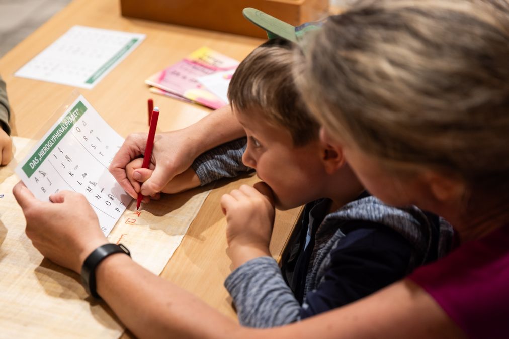 zur Vergrößerungsansicht des Bildes: Kind und Erwachsene mit einem Zettel auf dem der Name in Hierglyphen dargestellt ist.