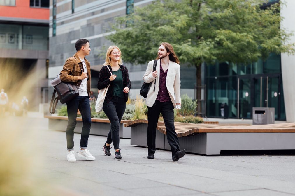 zur Vergrößerungsansicht des Bildes: Drei Studierende laufen über den Campus Augustusplatz und reden miteinander, im Vordergrund ist ein Grasbüchel verschwommen zu sehen, Foto: Christian Hüller