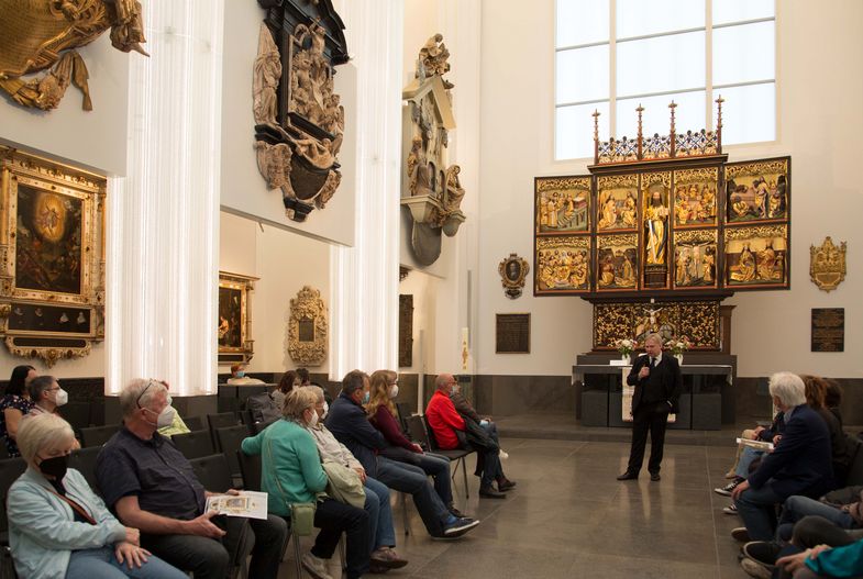 Blick in das Paulinum (Aula und Universitätskirche St. Pauli), vor dem Altar steht eine Person, die Erklärungen zu den Kunstwerken abgibt, die Gruppe, die an der Führung teilnimmt, sitzt auf Stühlen im Raum verteilt, an den Wänden sind die Epitaphien zu sehen