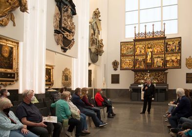 Blick in das Paulinum (Aula und Universitätskirche St. Pauli), vor dem Altar steht eine Person, die Erklärungen zu den Kunstwerken abgibt, die Gruppe, die an der Führung teilnimmt, sitzt auf Stühlen im Raum verteilt, an den Wänden sind die Epitaphien zu sehen