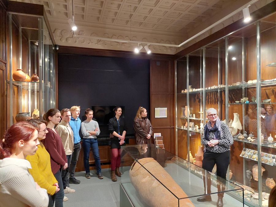 zur Vergrößerungsansicht des Bildes: Stipendiaten stehen in einem Raum des Ägyptischen Museums und hören der Kustodin zu den Ausfürungen zu, in der Mitte steht eine Glasvitrine mit einer Mumie