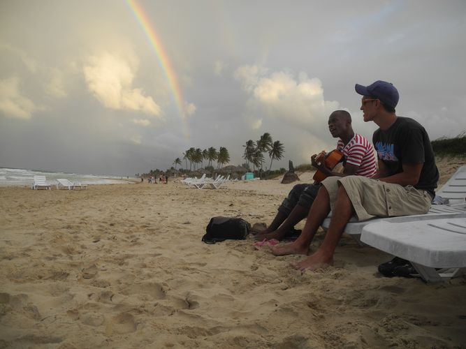 Das Bild zeigt zwei musizierende Männer am Strand. Im Hintergrund ist ein Regenbogen zu sehen.