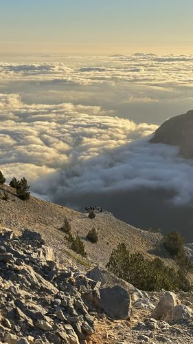 Blick vom Olymp in gleißendem Sonnenlicht in das wolkenverhangene Tal. Am Berghang in der Mitte des Bildes ist eine Wandergruppe zu sehen.