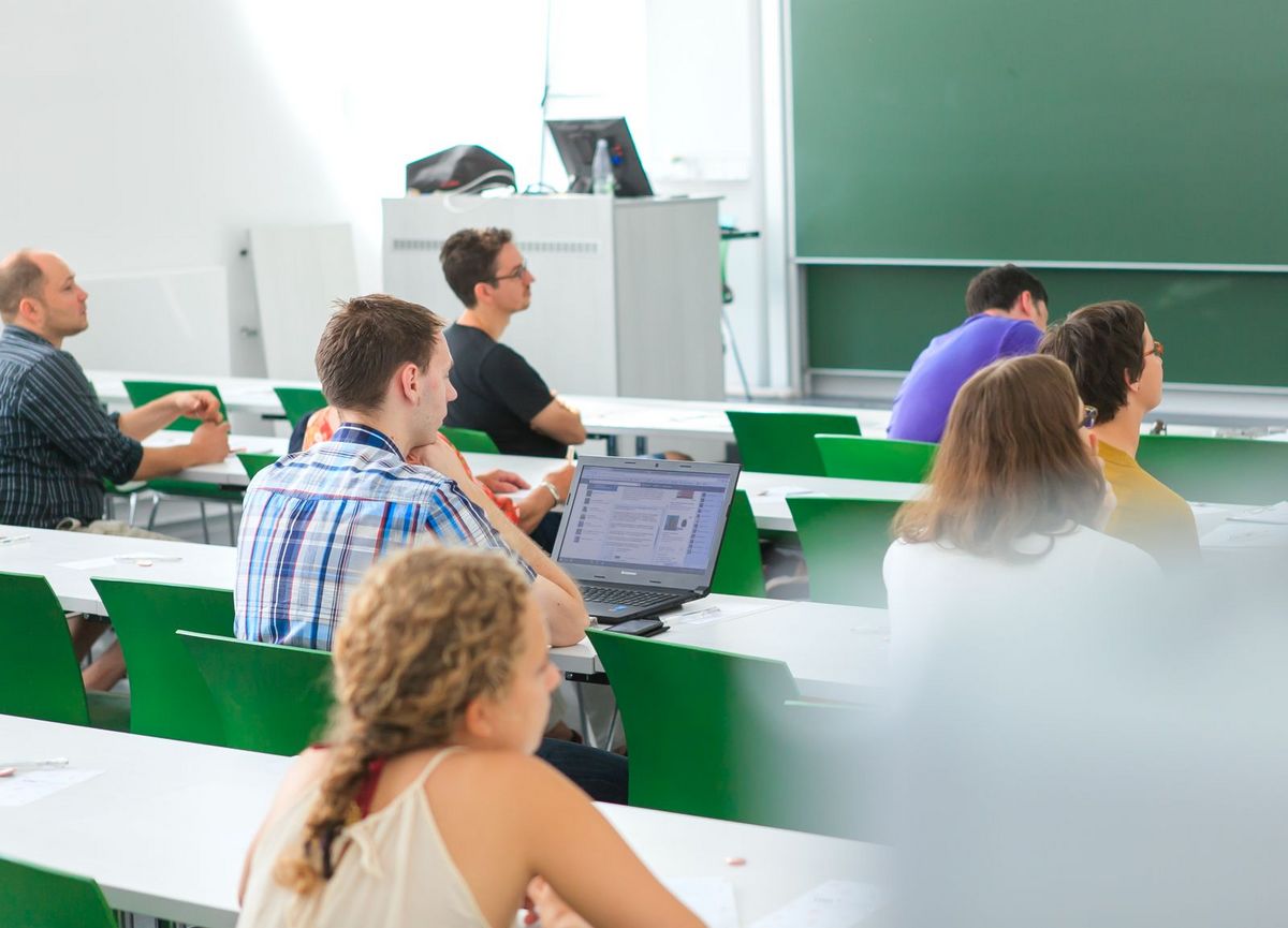 zur Vergrößerungsansicht des Bildes: Studierende sitzen im Hörsaal an Tischen verteilt und folgen einer Vorlesung