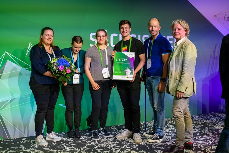 Zu sehen ist das Team von NEXT3D um Prof. Dr. Dirk Winkler und PD Dr. Ronny Grunert auf der Bühne, mit der Urkunde für den 2. Platz in der Hand.