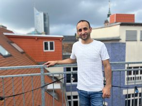 Mann steht auf Dachterasse bei Tageslicht und lächelt in die Kamera, im Hintergrund Häuser der Leipziger Innenstadt
